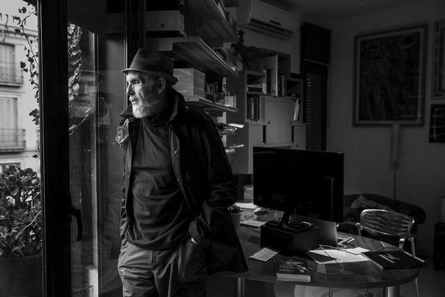 Ángel Alonso recolzat al brancal de la porta del seu estudi, amb prestatgeries i una taula de treball al fons